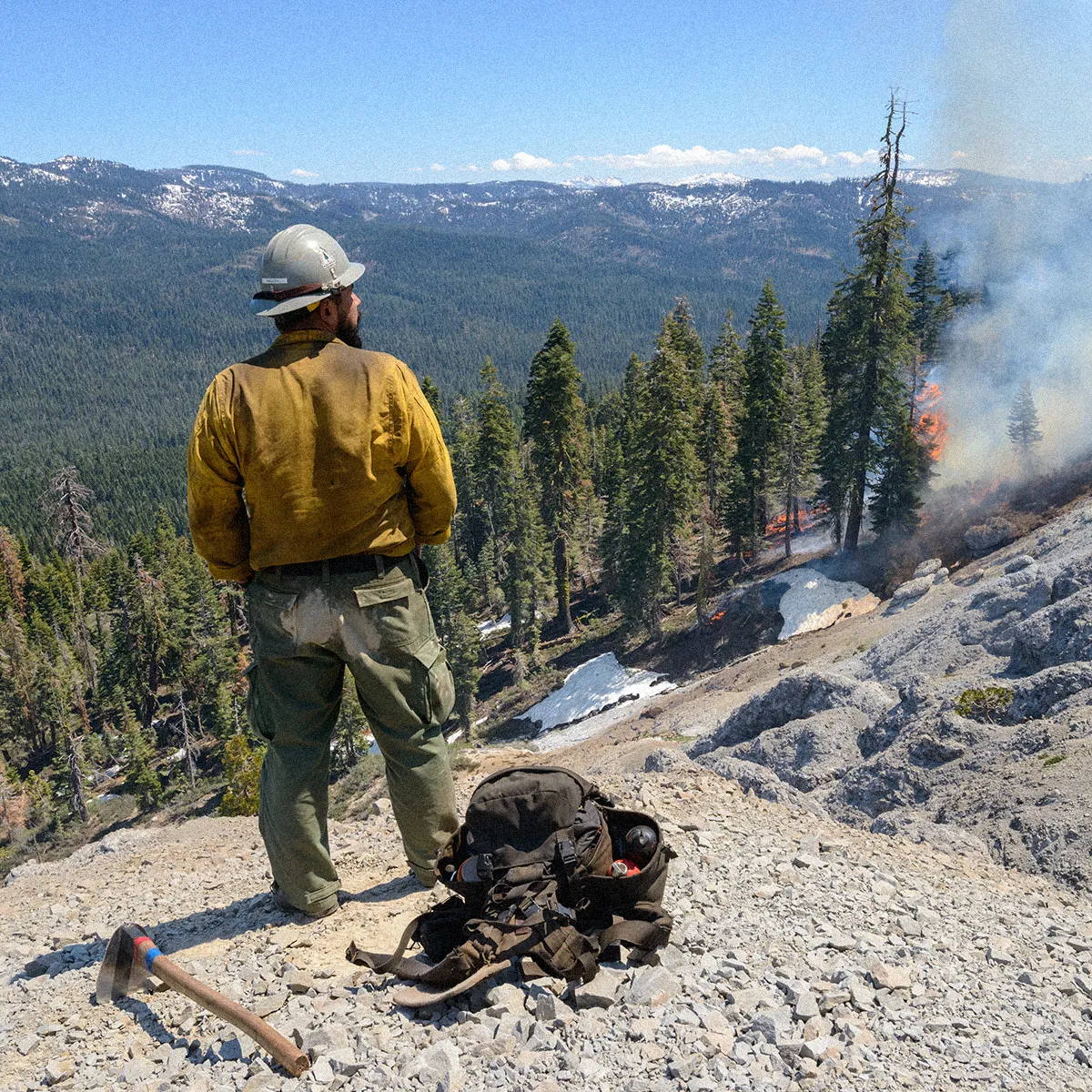 Wildfire crew overlooking mountain landscape from a rock pile, with a controlled burn occurring below.