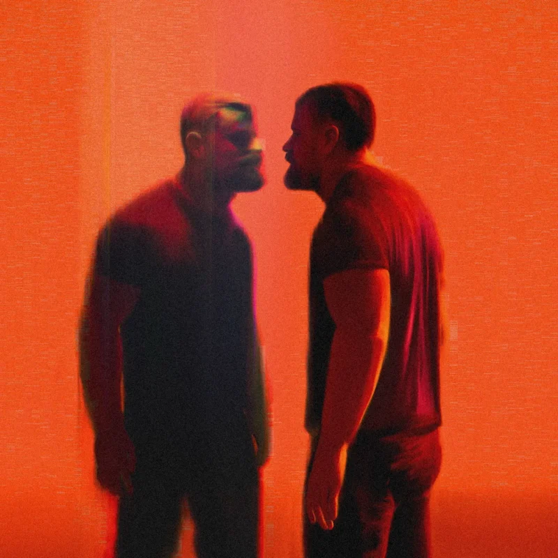 A burly man facing his reflection under red lighting backdrop.
