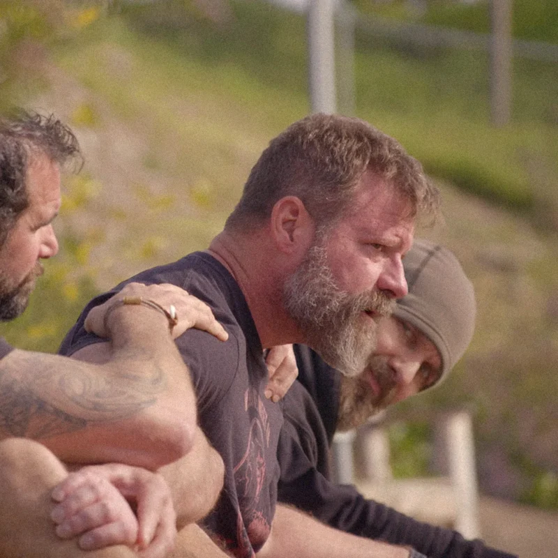 Group of three men resting outdoors, one comforting another by placing hands on shoulders.
